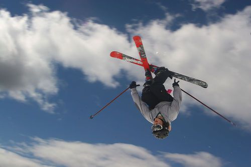 Alpine skiier crosses skis in the air during a jump at a ski mountain, clouds and sky in background.