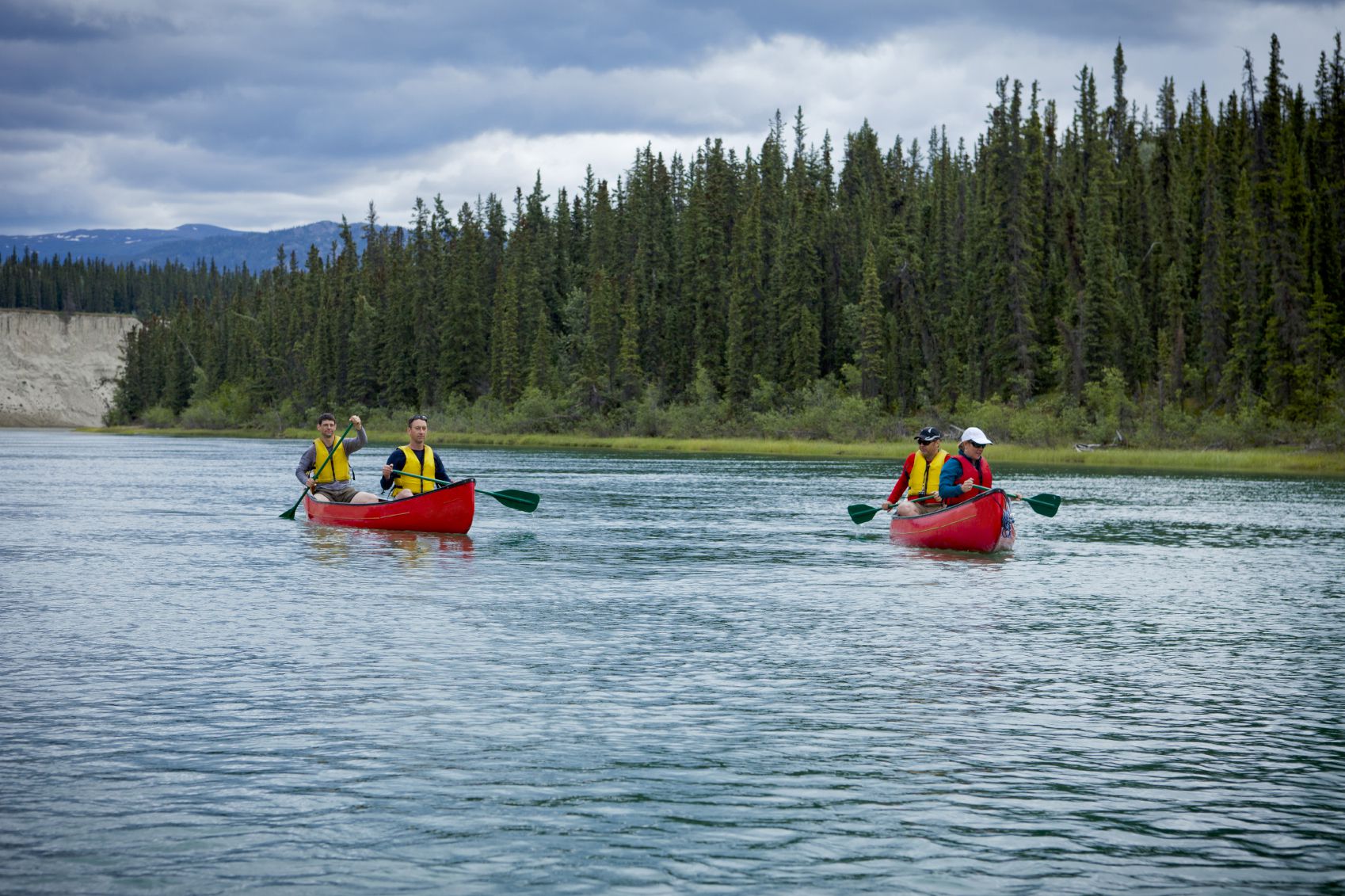 Two canoes that hold two people in each vessel coast down the blue Yukon River with Greenery and the blue sky behind them.